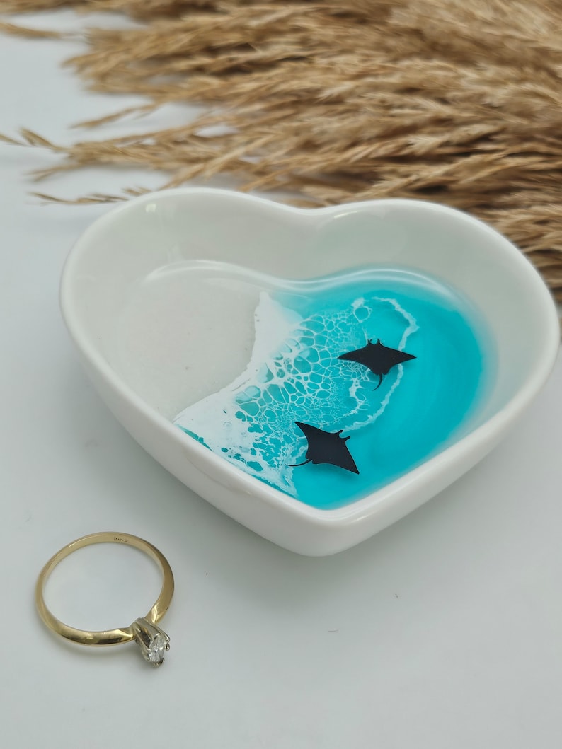 May include: A white heart-shaped trinket dish with a blue and white ocean-themed design. Two black manta ray silhouettes swim in the turquoise water. A gold ring with a diamond sits next to the dish.