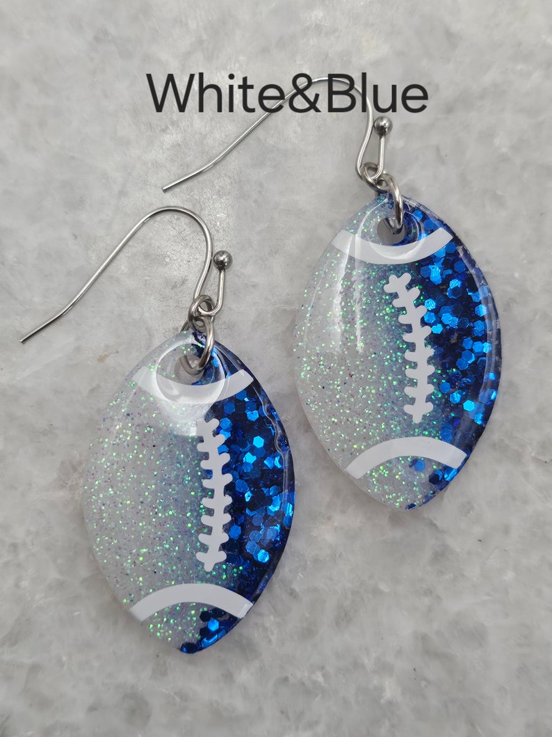 Op de afbeelding: Witte en blauwe glitter voetbal oorbellen. De oorbellen zijn gevormd als een voetbal en hebben witte glitter op de bovenste helft en blauwe glitter op de onderste helft. De voetbal veters zijn wit.