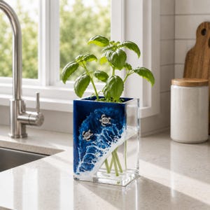 May include: A rectangular clear glass vase featuring a resin ocean wave design in shades of deep blue and white foam. Two small silver-toned sea turtle embellishments are attached to the front. A green basil plant is placed inside the vase.