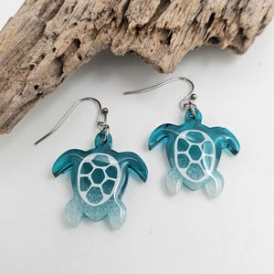May include: A pair of dangle earrings featuring sea turtle charms. The turtles are made of translucent teal resin with a white shell pattern and a gradient effect that fades into a white, glittery texture at the bottom of the flippers.