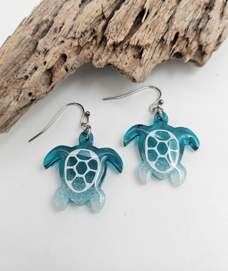 May include: A pair of dangle earrings featuring sea turtle charms. The turtles are made of translucent teal resin with a white shell pattern and a gradient effect that fades into a white, glittery texture at the bottom of the flippers.