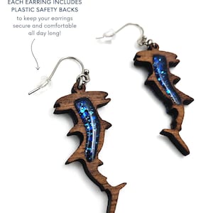 May include: A pair of hammerhead shark earrings crafted from dark brown wood with a blue glitter resin inlay. The earrings feature silver-toned metal hooks and include plastic safety backs. Text reads: EACH EARRING INCLUDES PLASTIC SAFETY BACKS.