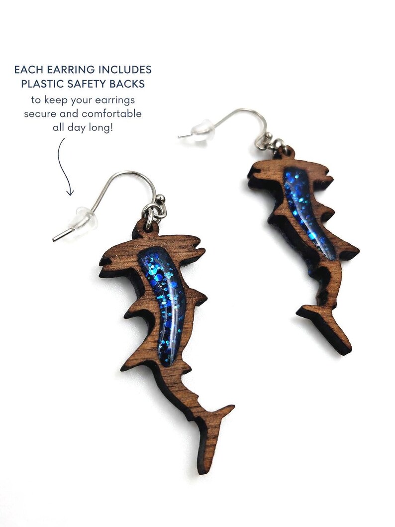 May include: A pair of hammerhead shark earrings crafted from dark brown wood with a blue glitter resin inlay. The earrings feature silver-toned metal hooks and include plastic safety backs. Text reads: EACH EARRING INCLUDES PLASTIC SAFETY BACKS.