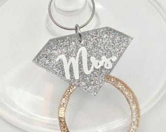 May include: A wine glass charm shaped like a diamond ring with a silver glitter diamond top featuring the white text Mrs. and a gold glitter band. The charm is attached to a thin silver metal hoop designed to hook onto the stem of a glass.