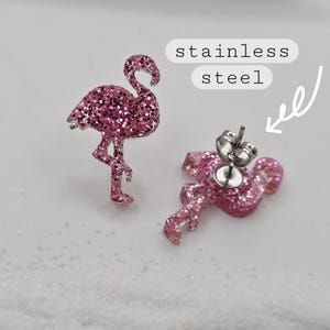 K&ouml;nnte beinhalten: Ein Paar Ohrstecker in Form von pinkfarbenen Glitzer-Flamingos. Ein Ohrstecker zeigt die Vorderseite, der andere die R&uuml;ckseite mit einem silberfarbenen Metallstift und Verschluss. Ein wei&szlig;es Etikett mit dem Text stainless steel zeigt auf den Stift.