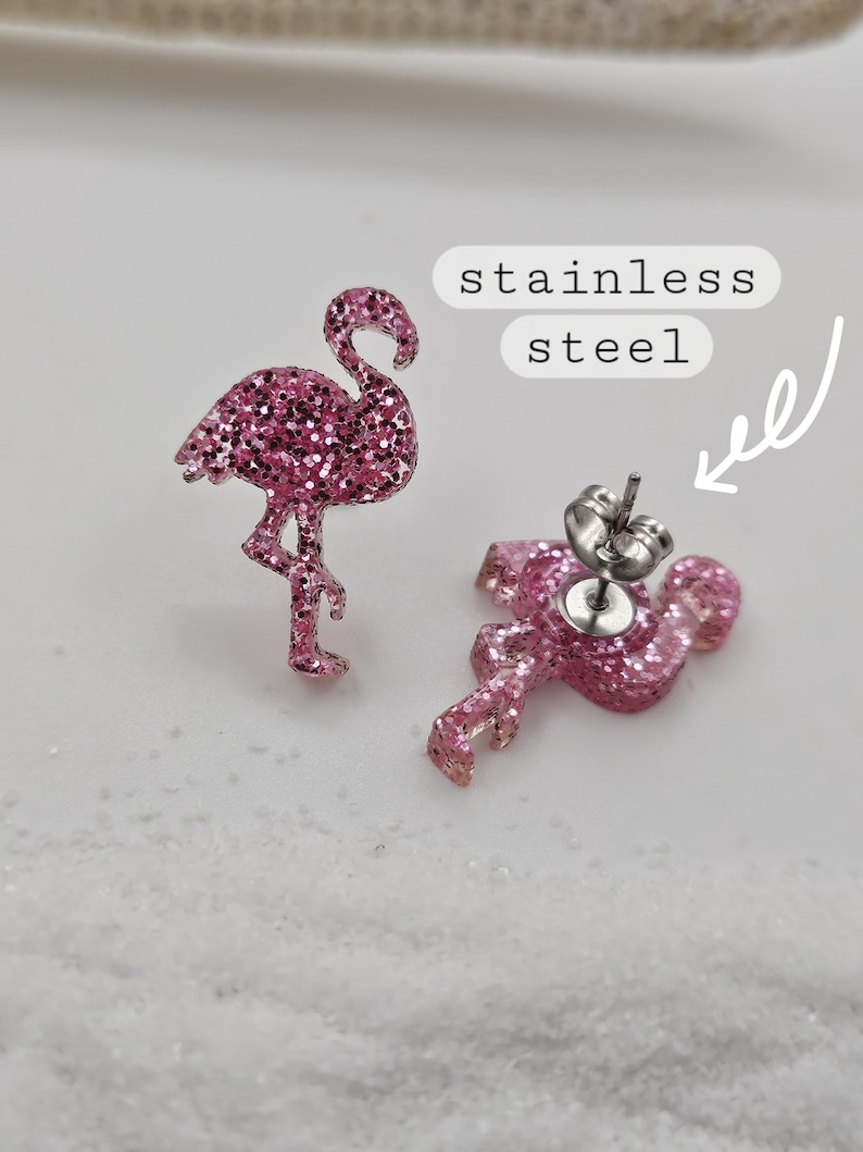 K&ouml;nnte beinhalten: Ein Paar Ohrstecker in Form von pinkfarbenen Glitzer-Flamingos. Ein Ohrstecker zeigt die Vorderseite, der andere die R&uuml;ckseite mit einem silberfarbenen Metallstift und Verschluss. Ein wei&szlig;es Etikett mit dem Text stainless steel zeigt auf den Stift.