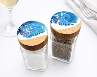 May include: Two square glass salt and pepper shakers with wooden lids. The lids feature a decorative blue and white resin pour design resembling ocean waves. One shaker contains white salt, and the other contains coarse black pepper.