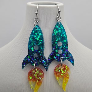May include: A pair of handmade rocket ship earrings. The earrings feature a teal glitter body with a dark blue glitter fin and a flame of orange and yellow glitter. The earrings hang from silver-colored ear wires.