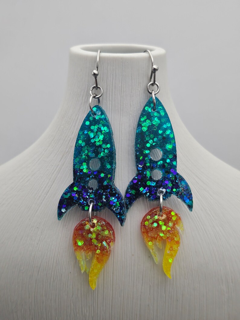 May include: A pair of handmade rocket ship earrings. The earrings feature a teal glitter body with a dark blue glitter fin and a flame of orange and yellow glitter. The earrings hang from silver-colored ear wires.