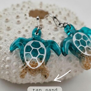 May include: A pair of dangle earrings featuring sea turtle charms. The turtles have translucent teal bodies, white shell patterns, and tan sand textures on the lower portions. A white arrow points to the sand, with the text tan sand below.