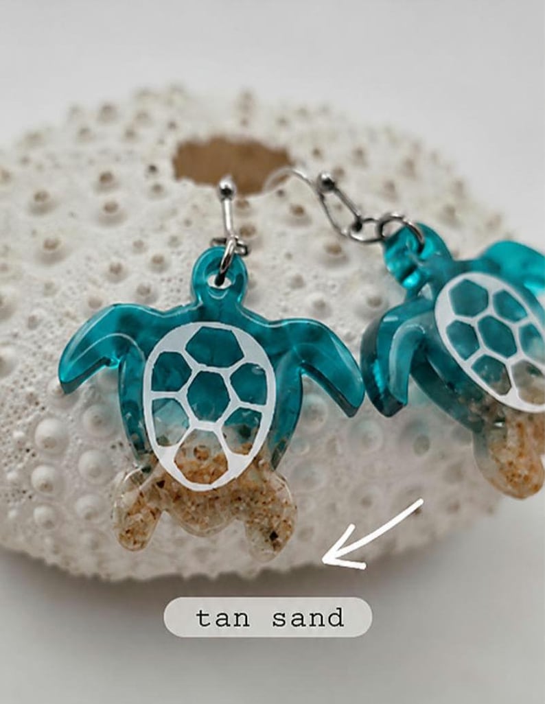 May include: A pair of dangle earrings featuring sea turtle charms. The turtles have translucent teal bodies, white shell patterns, and tan sand textures on the lower portions. A white arrow points to the sand, with the text tan sand below.