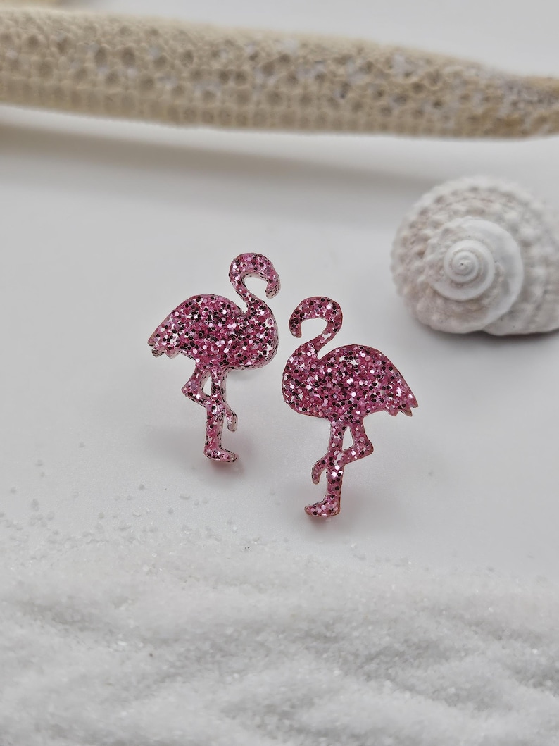 K&ouml;nnte beinhalten: Ein Paar Ohrstecker in Form von Flamingos mit einer leuchtend pinken Glitzerbeschichtung. Jeder Flamingo ist auf einem Bein stehend mit nach unten gebogenem Hals dargestellt und weist eine texturierte, funkelnde Oberfl&auml;che auf.