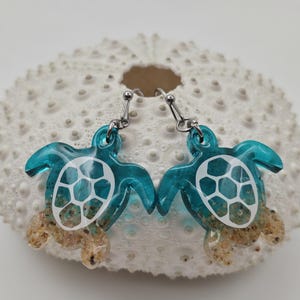 May include: A pair of dangle earrings featuring sea turtle charms made of translucent teal resin. The turtle shells have white geometric patterns, and the lower portions of the bodies are filled with tan sand. The earrings hang from silver-toned metal hooks.