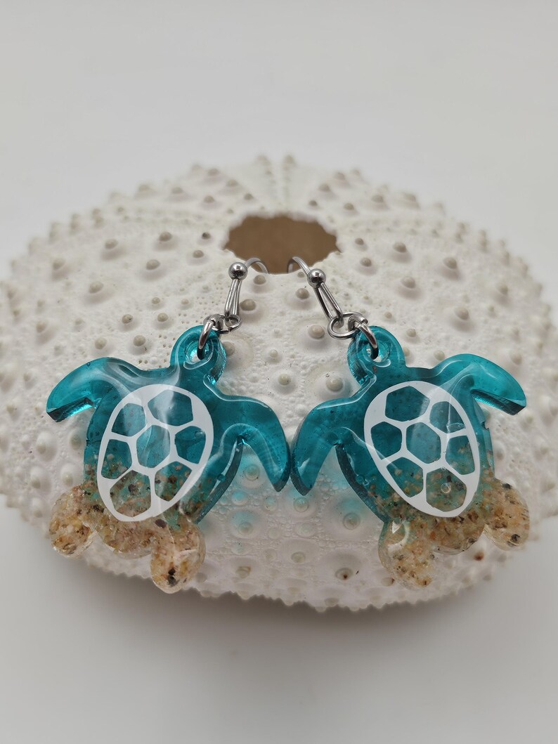 May include: A pair of dangle earrings featuring sea turtle charms made of translucent teal resin. The turtle shells have white geometric patterns, and the lower portions of the bodies are filled with tan sand. The earrings hang from silver-toned metal hooks.