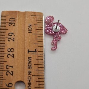 K&ouml;nnte beinhalten: Ein rosa glitzernder Flamingo-Ohrstecker mit Metallstift, platziert neben einem Holzlineal, das zeigt, dass der Flamingo eine H&ouml;he von etwa 2,5 cm vom Kopf bis zum unteren Ende des Beins aufweist.