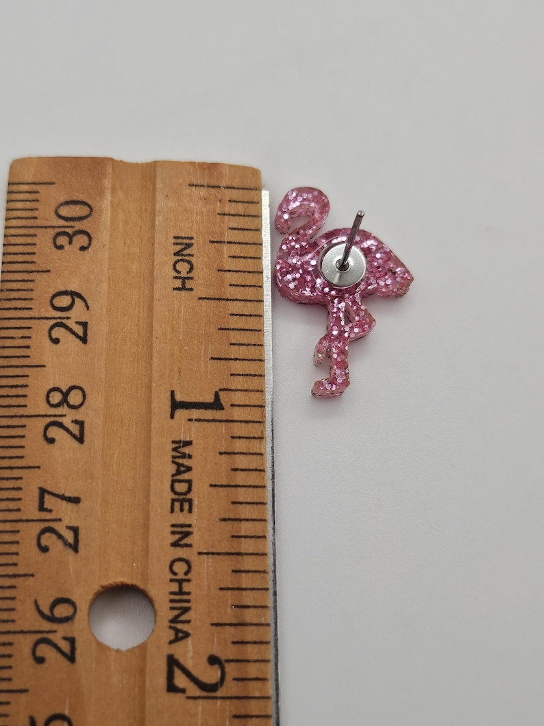 K&ouml;nnte beinhalten: Ein rosa glitzernder Flamingo-Ohrstecker mit Metallstift, platziert neben einem Holzlineal, das zeigt, dass der Flamingo eine H&ouml;he von etwa 2,5 cm vom Kopf bis zum unteren Ende des Beins aufweist.