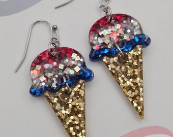 May include: A pair of dangle earrings shaped like ice cream cones. The scoops are made of red, white, and blue glitter resin, while the cones are filled with gold glitter. Each earring features a silver-toned metal hook for attachment.