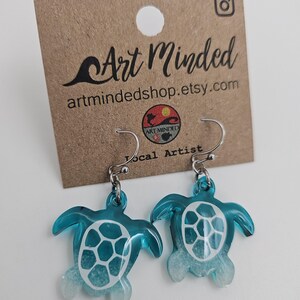 May include: A pair of dangle earrings featuring translucent teal sea turtle charms with white shell patterns and white-tipped flippers. The earrings are attached to a brown cardboard display card printed with the text Art Minded and artmindedshop.etsy.com.