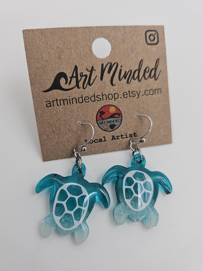 May include: A pair of dangle earrings featuring translucent teal sea turtle charms with white shell patterns and white-tipped flippers. The earrings are attached to a brown cardboard display card printed with the text Art Minded and artmindedshop.etsy.com.