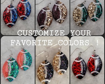 Pode incluir: Seis pares de brincos em forma de bola de futebol com glitter e costuras brancas. Cada par apresenta uma combinação de cores diferente, incluindo vermelho e azul, bordô e dourado, dourado e verde, laranja e turquesa, dourado e preto e vermelho e preto.