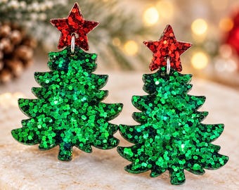Puede incluir: Un par de pendientes colgantes con forma de árbol de Navidad. Cada pendiente presenta un cuerpo de árbol verde con purpurina y una pequeña estrella roja brillante sujeta en la parte superior mediante una anilla de metal plateado festiva.
