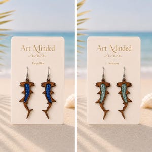 May include: Two pairs of hammerhead shark earrings on display cards. One pair features dark wood with deep blue inlay, the other features dark wood with seafoam green inlay. Each earring has a silver-toned metal hook and a cutout shark silhouette.