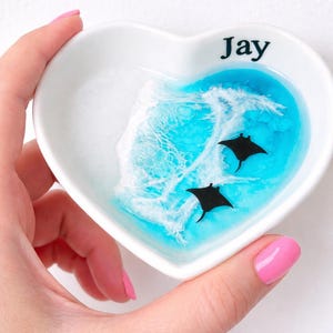 May include: Heart-shaped white ceramic trinket dish with a blue and white ocean wave design. Two black manta rays swim in the blue water. The name "Jay" is printed in black on the rim.