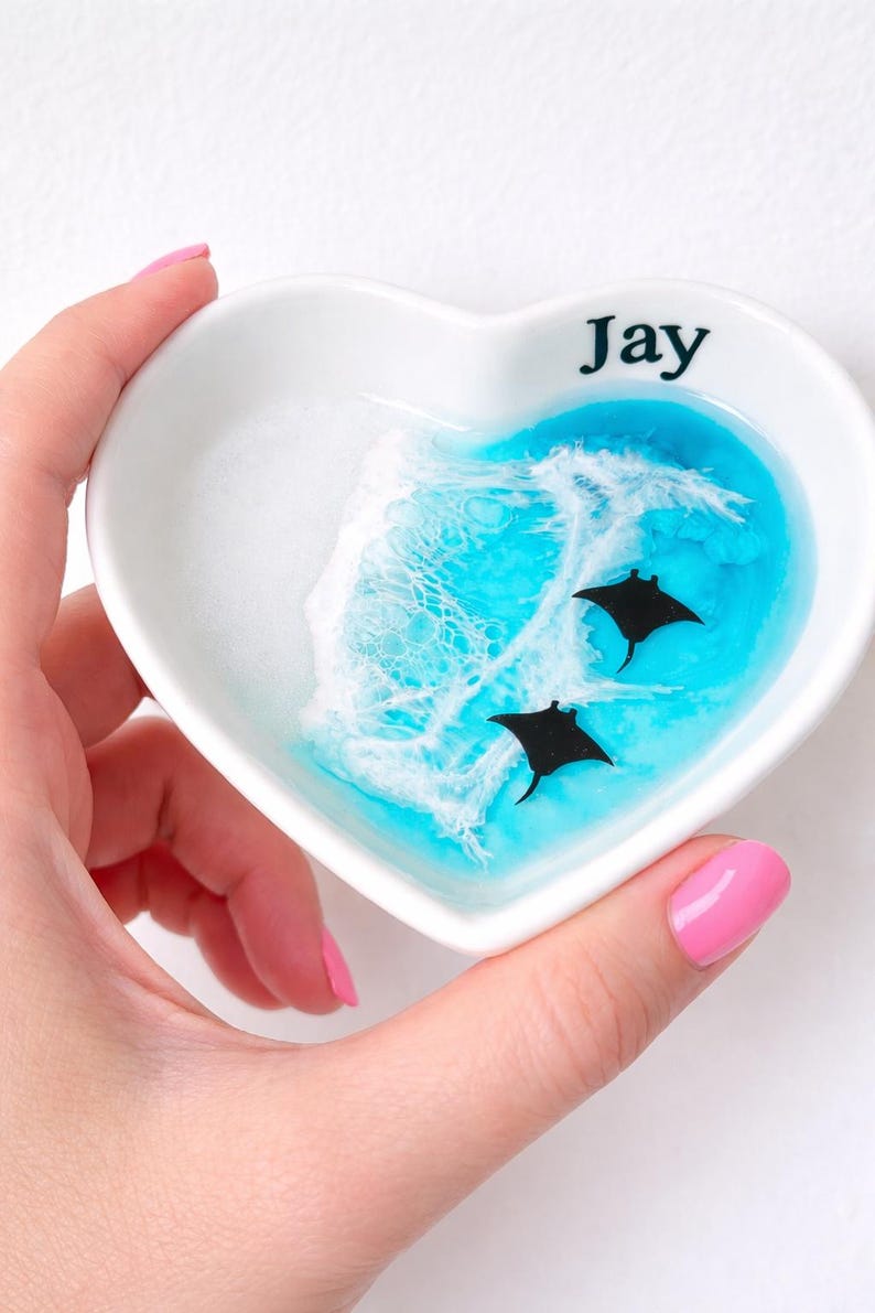 May include: Heart-shaped white ceramic trinket dish with a blue and white ocean wave design. Two black manta rays swim in the blue water. The name "Jay" is printed in black on the rim.