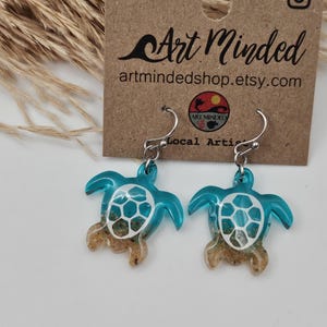 May include: A pair of sea turtle earrings featuring a translucent teal resin body with a white shell pattern and a sandy texture at the base. The earrings hang from silver-toned metal hooks and are displayed on a brown card labeled Art Minded.