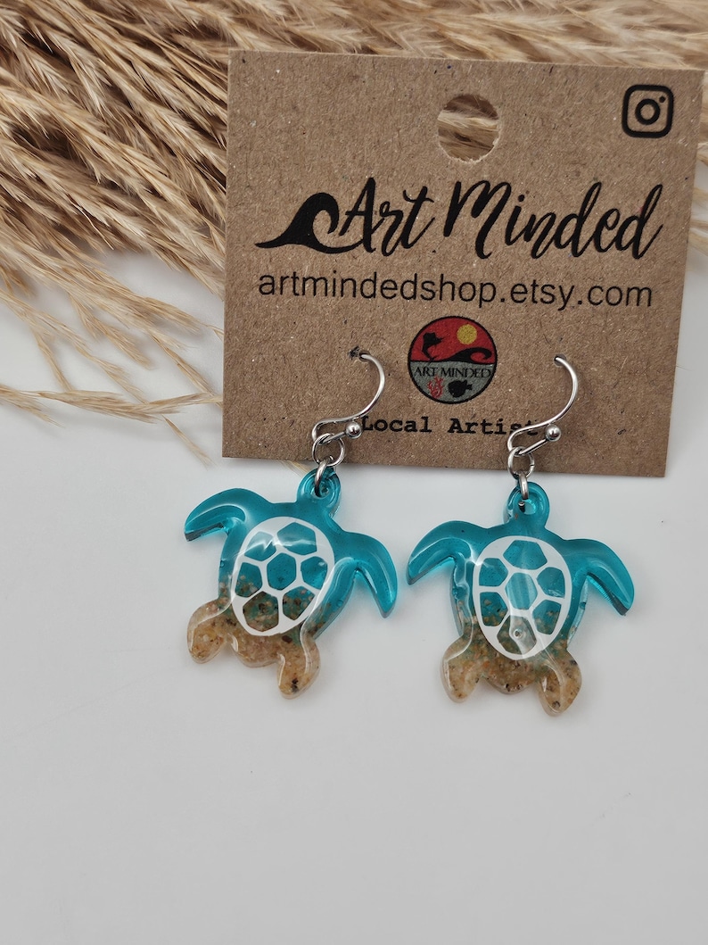 May include: A pair of sea turtle earrings featuring a translucent teal resin body with a white shell pattern and a sandy texture at the base. The earrings hang from silver-toned metal hooks and are displayed on a brown card labeled Art Minded.