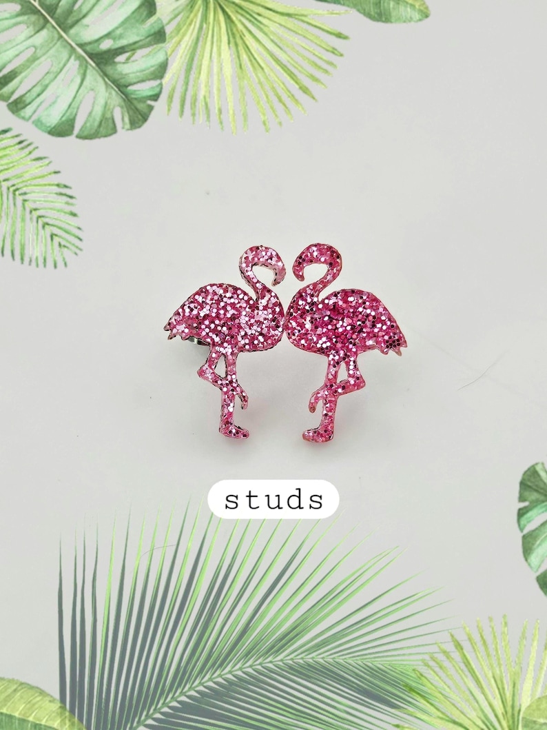 K&ouml;nnte beinhalten: Ein Paar Ohrstecker in Form von rosafarbenen Flamingos, die mit glitzernden Pailletten bedeckt sind. Die beiden Flamingos stehen sich gegen&uuml;ber und das Wort studs steht in einem wei&szlig;en Rechteck unter den Ohrringen, umgeben von Palmenbl&auml;ttern.