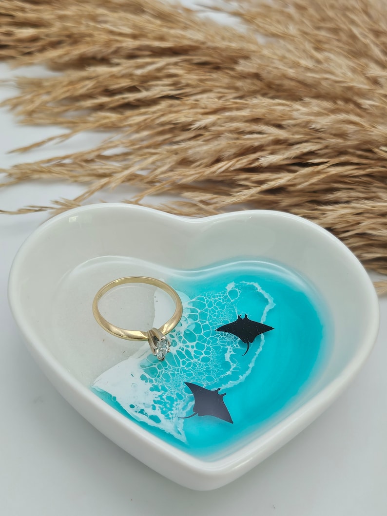 May include: A heart-shaped white trinket dish with a turquoise and white resin design, featuring two black manta ray silhouettes. A gold ring with a clear gemstone sits in the dish. Decorative dried grass is in the background.