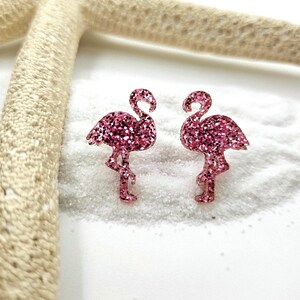 K&ouml;nnte beinhalten: Ein Paar Ohrstecker in Form von Flamingos mit einer leuchtend pinkfarbenen Glitzerbeschichtung. Jeder Flamingo ist in stehender Pose mit gebogenem Hals dargestellt und weist eine texturierte, funkelnde Oberfl&auml;che auf, die das Licht reflektiert.