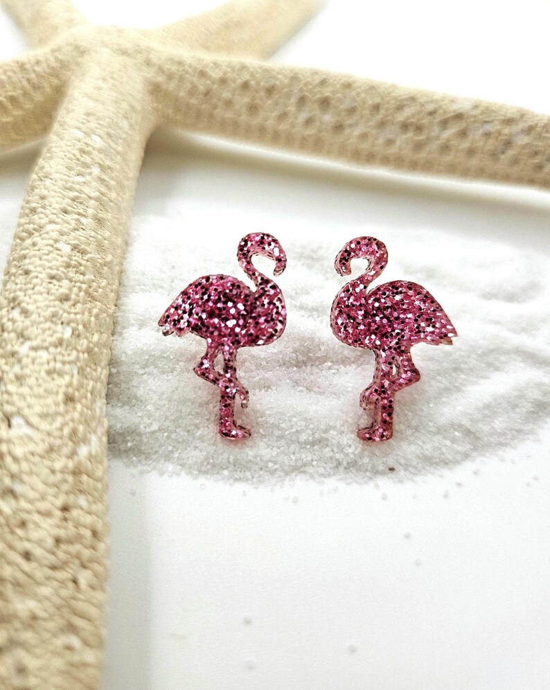 K&ouml;nnte beinhalten: Ein Paar Ohrstecker in Form von Flamingos mit einer leuchtend pinkfarbenen Glitzerbeschichtung. Jeder Flamingo ist in stehender Pose mit gebogenem Hals dargestellt und weist eine texturierte, funkelnde Oberfl&auml;che auf, die das Licht reflektiert.