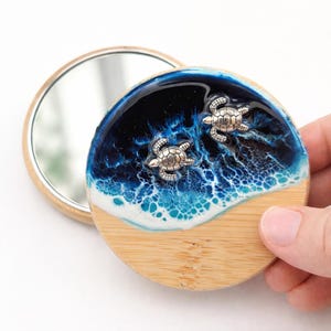 May include: A round, open compact mirror with a bamboo base. The compact features a resin design of a blue ocean with white waves and two silver sea turtles. The mirror is open, revealing the reflective surface.