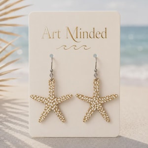 May include: A pair of gold and white starfish earrings on a cream-colored display card with the text Art Minded printed in gold. The earrings feature a textured, speckled surface and hang from silver-toned metal hooks on the rectangular card.