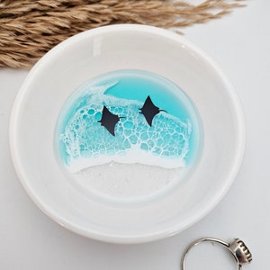 May include: A white ceramic ring dish with a turquoise and white resin design featuring two black manta ray silhouettes. A silver ring with a dark stone sits next to the dish. Dried wheat stalks are in the background.