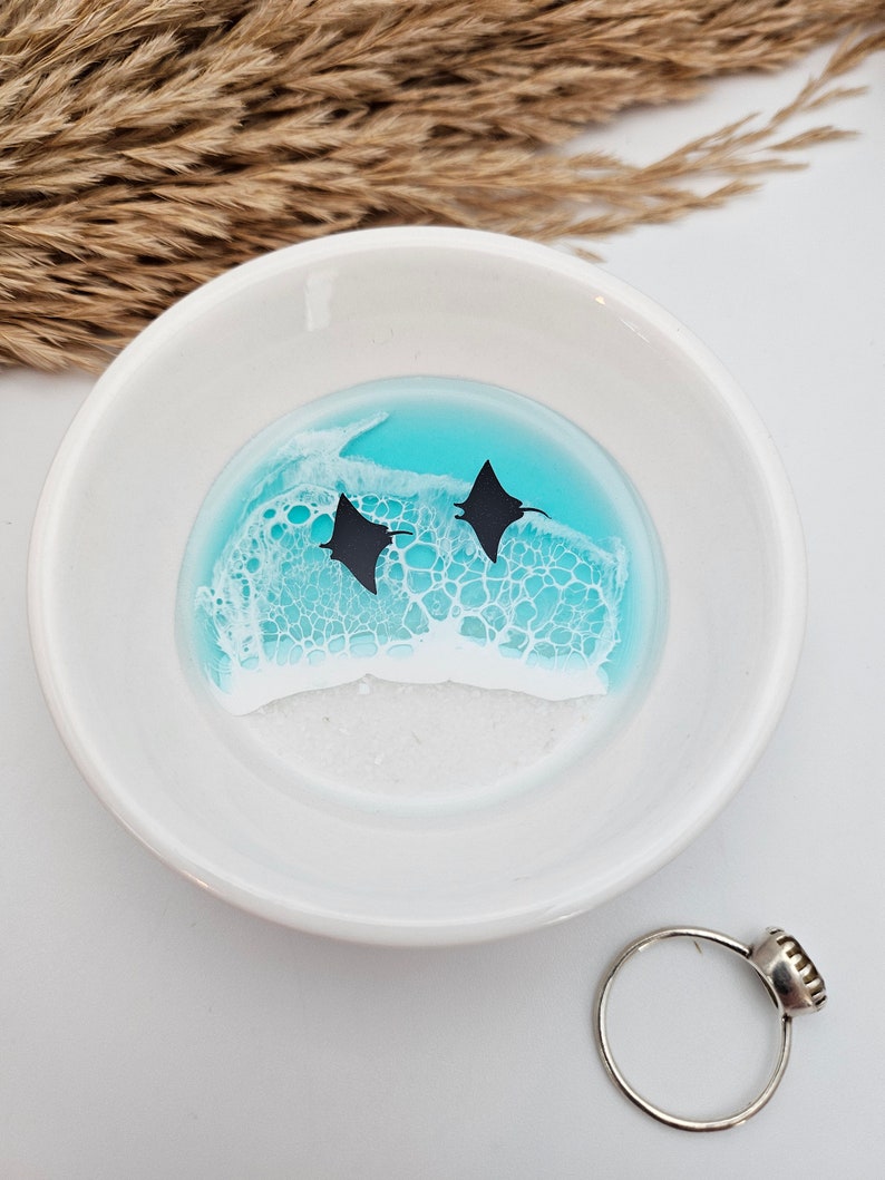 May include: A white ceramic ring dish with a turquoise and white resin design featuring two black manta ray silhouettes. A silver ring with a dark stone sits next to the dish. Dried wheat stalks are in the background.