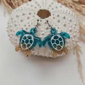May include: A pair of dangle earrings shaped like sea turtles. The turtle shells are made of translucent teal resin with a white geometric pattern, while the lower portions of the bodies are filled with tan sand and clear resin. Silver-toned hooks.