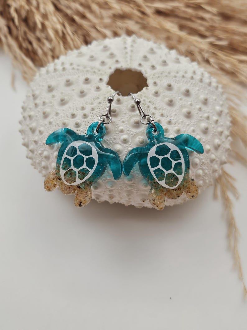 May include: A pair of dangle earrings shaped like sea turtles. The turtle shells are made of translucent teal resin with a white geometric pattern, while the lower portions of the bodies are filled with tan sand and clear resin. Silver-toned hooks.