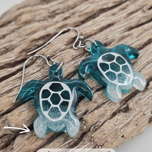 May include: A pair of dangle earrings featuring translucent teal sea turtle charms with white sand detailing at the base of the flippers and a white shell pattern on the back. The earrings include silver-toned metal hooks for attachment.