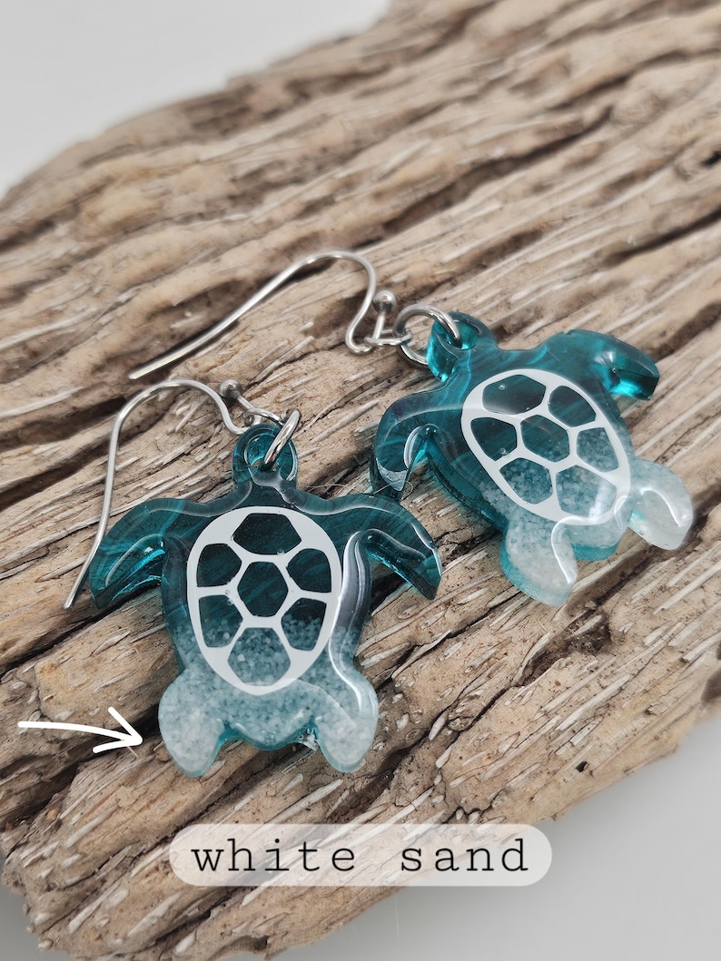 May include: A pair of dangle earrings featuring translucent teal sea turtle charms with white sand detailing at the base of the flippers and a white shell pattern on the back. The earrings include silver-toned metal hooks for attachment.