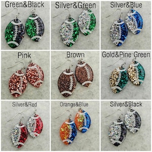 Op de afbeelding: Negen paar voetbalvormige oorbellen met glitter en verschillende kleurcombinaties. De kleuren omvatten groen, zwart, zilver, blauw, roze, bruin, goud, pijnboomgroen, rood, oranje en wit.