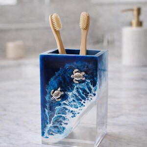 May include: A rectangular toothbrush holder featuring a deep blue and white ocean wave design with two silver-toned sea turtle embellishments on the front. The holder is made of clear resin and contains two light-colored wooden toothbrushes.