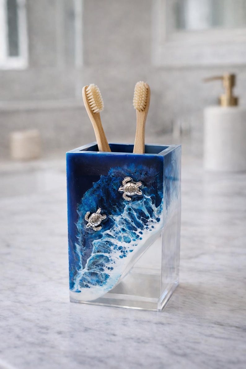 May include: A rectangular toothbrush holder featuring a deep blue and white ocean wave design with two silver-toned sea turtle embellishments on the front. The holder is made of clear resin and contains two light-colored wooden toothbrushes.