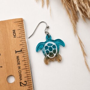 May include: A sea turtle shaped earring with a silver-toned hook. The turtle features a translucent blue head and flippers, a white shell pattern, and a sandy-textured base. A wooden ruler is visible, showing the earring is approximately 1.25 inches.