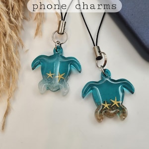 May include: Two teal turtle phone charms with gold starfish accents and a clear base. The charms are attached to black cords with silver-colored metal clasps. The text "phone charms" is in a white rectangle.