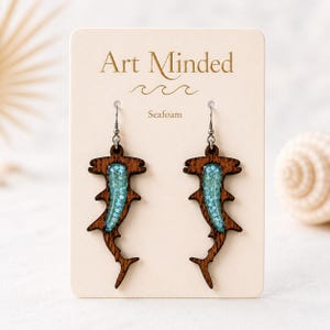 May include: A pair of hammerhead shark earrings made of dark brown wood with a central inlay of light blue glitter resin. The earrings are attached to a cream-colored display card labeled Art Minded and Seafoam with a wave graphic.