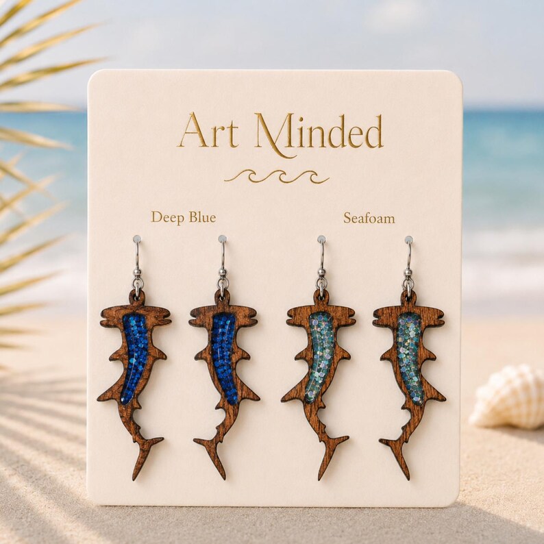 May include: Two pairs of hammerhead shark-shaped earrings on a display card labeled Art Minded. One pair features deep blue glitter inlay, and the other pair features seafoam green glitter inlay. The earrings are made of brown wood with metal hooks.