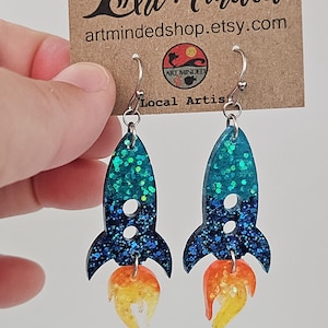 May include: A pair of handmade rocket ship earrings. The earrings feature a teal and blue glitter design with orange and yellow flames. The earrings are hanging from silver hooks. The background includes a brown tag with the text "Art Minded" and "artmindedshop.etsy.com."